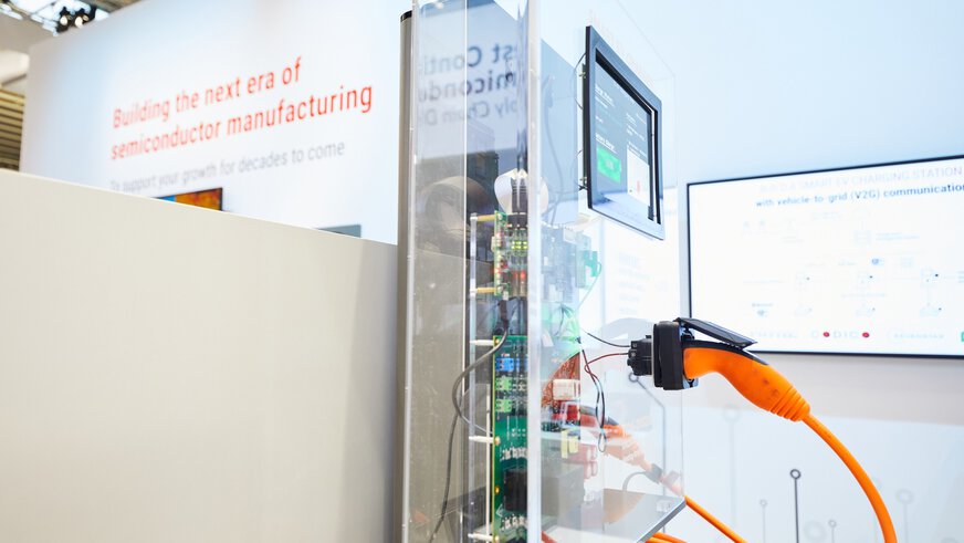 Ein oranges Ladekabel ist an eine Ladekonsole aus Plexiglas angeschlossen. Ein Display zeigt Informationen an und im Inneren ist die Elektronik zu sehen.