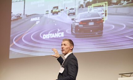 Ein Mann im Anzug präsentiert auf einer Bühne, die Leinwand zeigt ein Autos auf einer Straße mit digitalen Überlagerungen für Winkel- und Entfernungsmessungen.