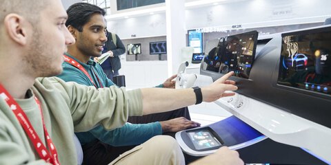 Zwei Personen sitzen vor einem, an einem Messestand ausgestellten, futuristsichen Amaturenbrett und interagieren mit der Technologie.