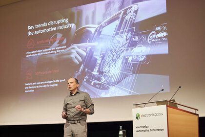 Ein Mann spricht auf der Bühne der electronica. Er steht neben dem Podium und hinter ihm ist eine Folie über die automotive industry auf der Leinwand zu sehen.